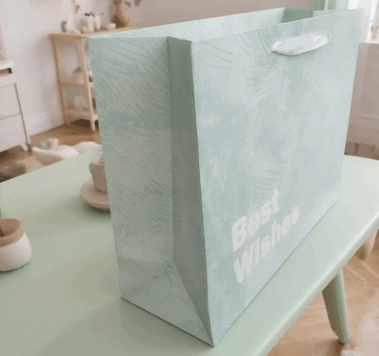 This handbag is named FH-22019-L Cross-border Wholesale Simple Cardstock Gift Bag with Good Wishes. It is made of high-quality cardstock material, which is crisp and well-shaped, and can firmly hold items. The surface is printed with fine techniques, with a light blue tone that is fresh and elegant. Paired with simple text patterns, it is simple yet stylish. The white handle design is in harmony with the color of the bag body and is also sturdy and durable. It is suitable for carrying goods and gift packaging when shopping, or for daily storage and carrying small items, etc. Whether it is for shopping or gift-giving occasions, it can show elegant taste.Product uses: Packaging paper bags, shopping paper bags, gift paper bags, festival paper bags, promotional paper bags.